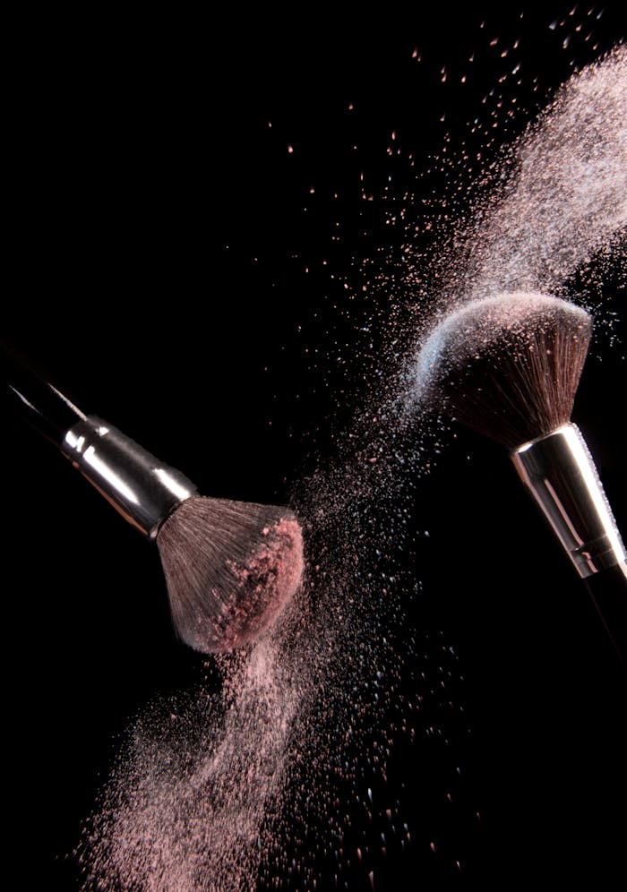 Home High-speed capture of makeup brushes dispersing pink powder in motion against a dark background.